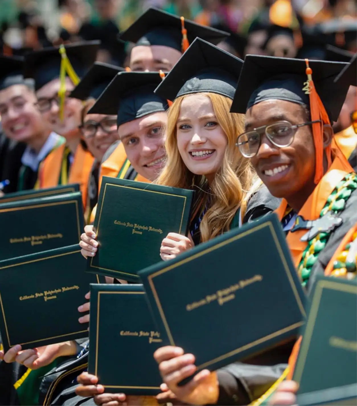 A group of graduate students dressed in black, green and gold robes, smiling as they hold their diplomas during their graduation ceremony.