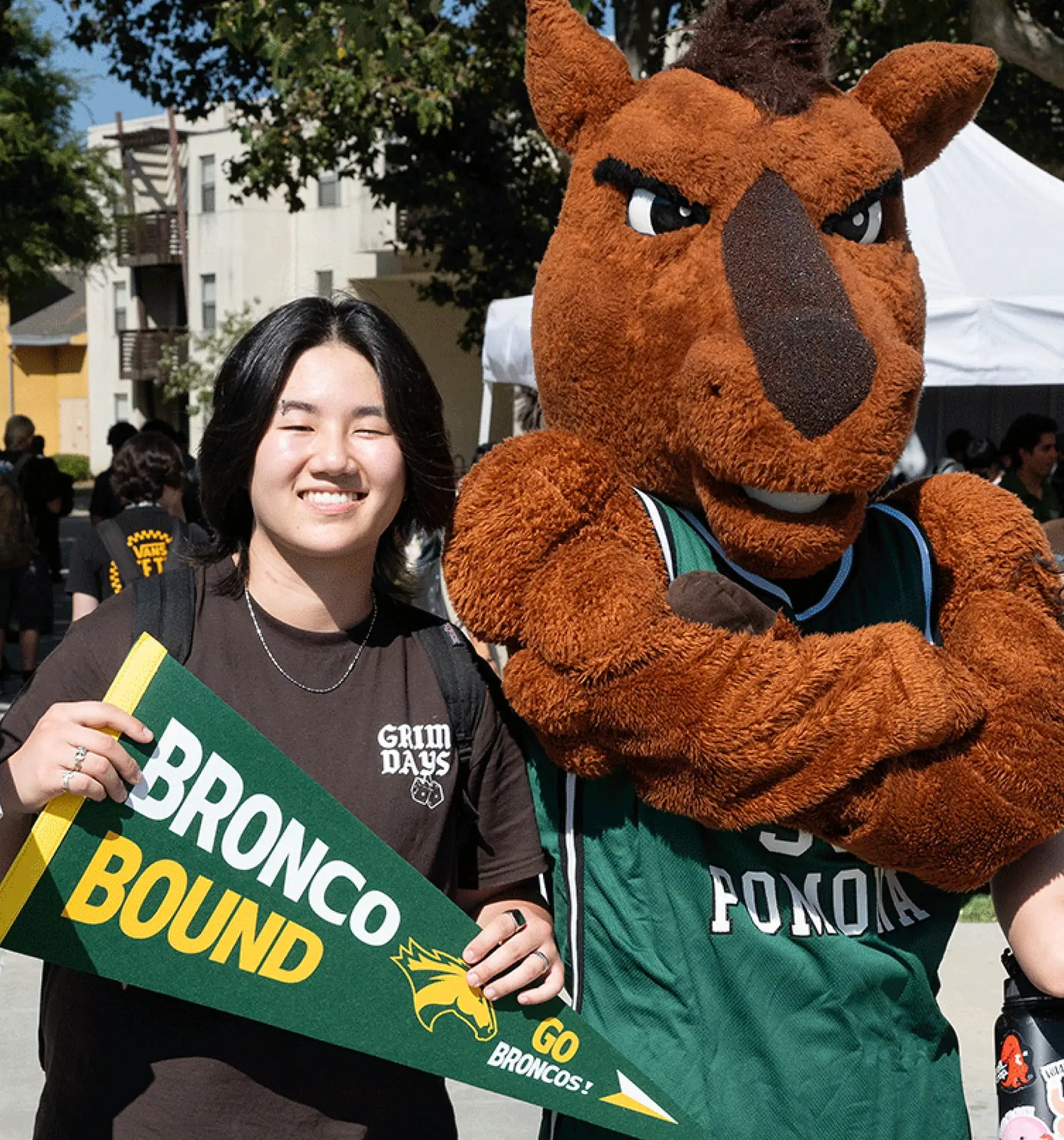 A female student smiles proudly while holding a sign in front of her as she stands next to the Cal Poly Pomona mascot.